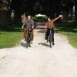 A couple rides bicycles along a path surrounded by green vineyards in Saint-Émilion, with rolling hills and historic stone buildings in the background under a sunny sky.